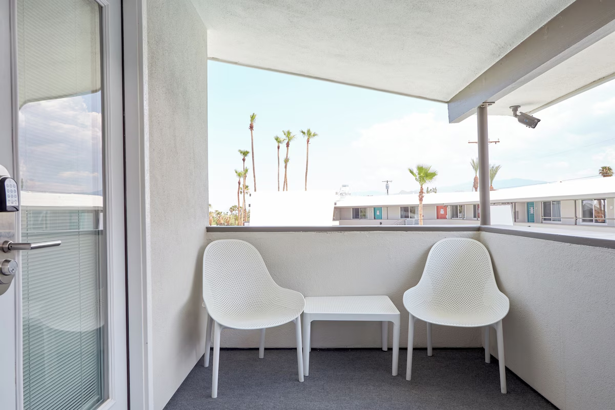 A small balcony with two white chairs and a small table, overlooking a courtyard with palm trees.