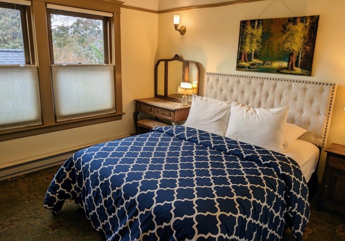 A neatly made bed with a blue and white comforter and white pillows in a cozy guest room with a window, painting, and nightstand.